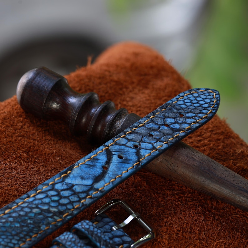 May include: A close-up of a blue watch strap with a textured pattern and orange stitching. The strap is made of leather and has a silver buckle. A dark wooden tool is in the background.