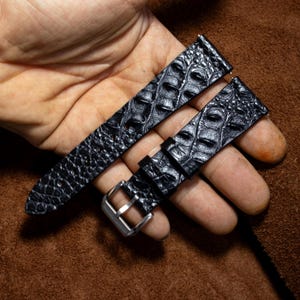 May include: A black watch strap featuring a textured crocodile skin pattern. The strap includes a silver-toned metal buckle and two matching leather keepers. The surface of the strap has a glossy finish with distinct, raised scales throughout.