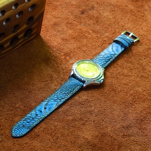 May include: A wristwatch with a blue leather band featuring a scale-like texture and yellow stitching. The watch face is silver with a yellow dial. The watch is set against a brown leather surface.
