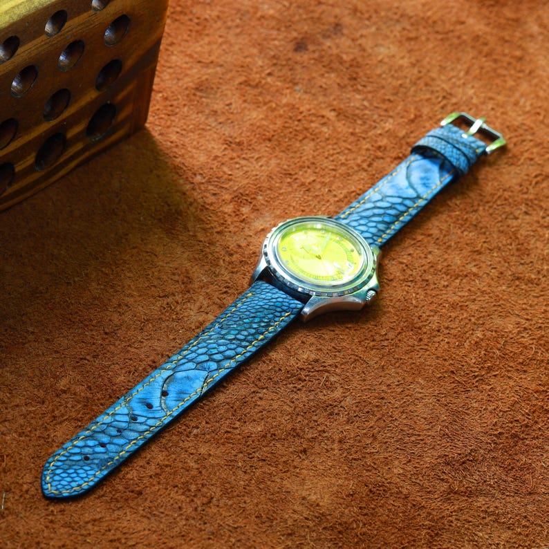 May include: A wristwatch with a blue leather band featuring a scale-like texture and yellow stitching. The watch face is silver with a yellow dial. The watch is set against a brown leather surface.