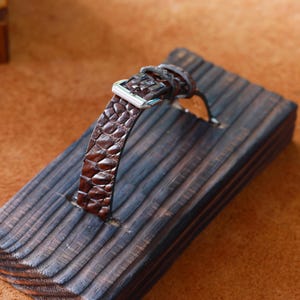 Op de afbeelding: Een bruine leren horlogeband met krokodillenpatroon wordt getoond op een donker houten blok. De band heeft een zilveren gesp. Het houten blok heeft een groef om de band vast te houden. De achtergrond is warm bruin.