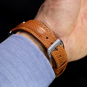 May include: A tan watch strap with a textured, lizard-like pattern and visible yellow stitching. The strap features a polished silver-tone metal buckle and is secured around a wrist, partially covered by a light blue, fine-striped long-sleeve shirt.