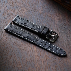 Puede incluir: Correa de reloj de cuero negro con costuras blancas. La correa presenta una hebilla plateada y está diseñada para un reloj clásico. El cuero tiene una apariencia texturizada, lo que se suma a su estética refinada.