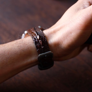 Könnte beinhalten: Nahaufnahme eines braunen Leder-Uhrenarmbands mit einem strukturierten, reptilienartigen Muster. Das Armband wird mit einer silberfarbenen Schnalle befestigt. Das Uhrenarmband ist um ein Handgelenk gelegt, was das Accessoire hervorhebt.