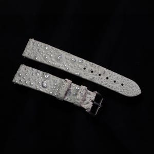 Puede incluir: Una correa de reloj de dos piezas en color blanco roto claro con una superficie texturizada y en relieve que recuerda a la piel de reptil. La correa presenta detalles metálicos plateados incrustados y una hebilla de metal plateado pulido.