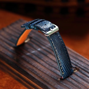 Peut inclure: Gros plan sur un bracelet de montre bleu avec un motif texturé et une boucle argentée. Le bracelet a une doublure orange et des coutures de couleur claire. Le bracelet de montre est présenté sur une surface en bois.