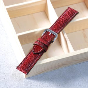 Könnte beinhalten: Ein rotes Lederarmband mit strukturierter Oberfläche und weißen Nähten. Das Armband hat eine silberfarbene Schnalle und wird auf einer hellen Holzoberfläche präsentiert. Das Armband ist etwa 23 cm lang.