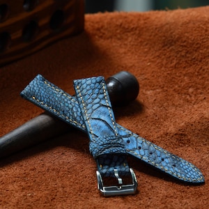May include: Blue watch strap with a textured, patterned surface. The strap features yellow stitching and a silver buckle. The leather strap is displayed on a brown surface.