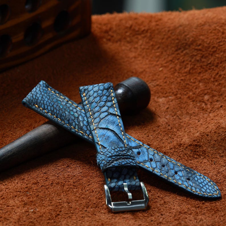 May include: Blue watch strap with a textured, patterned surface. The strap features yellow stitching and a silver buckle. The leather strap is displayed on a brown surface.