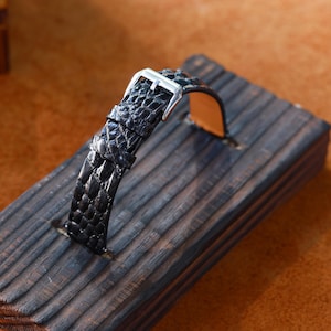 May include: A black watch strap with a textured, crocodile-like pattern. The strap has a silver-coloured buckle and is displayed on a dark wooden block. The watch band is likely made of leather.