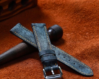 May include: Two watch straps made of distressed blue leather with yellow stitching. The straps have a silver-colored buckle and are lying on a brown surface. The watch straps are a stylish accessory.