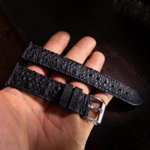 Pode incluir: Uma pulseira de relógio preta com um padrão texturizado em relevo que lembra pele de réptil. A pulseira inclui uma fivela de metal prateado e dois passantes pretos a condizer para prender a banda. A superfície tem um acabamento mate.