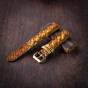 Puede incluir: Correa de reloj de dos piezas con un patrón texturizado similar al de un reptil en tonos marrones y dorados. La correa tiene una hebilla dorada y se muestra sobre una superficie de madera oscura.