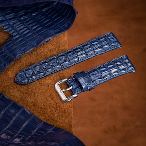May include: Two navy blue watch straps with a crocodile pattern. The straps have a silver buckle and are set against a brown leather background. The watch straps are made of a textured material.