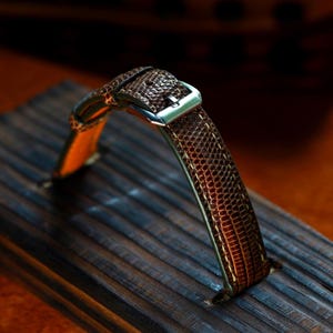 以下が含まれることがあります： 茶色の時計ベルト。爬虫類のような質感で、銀色のバックルが付いています。木製の表面に置かれています。ベルトの内側はオレンジ色です。
