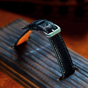 Puede incluir: Primer plano de una correa de reloj negra con un patrón texturizado y costuras blancas. La correa presenta una hebilla plateada y una parte inferior naranja. La correa del reloj se muestra sobre una superficie de madera.