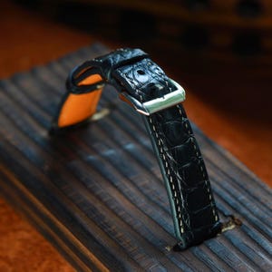 Peut inclure: Bracelet de montre noir avec une texture peau de crocodile, une boucle argentée et des coutures blanches. Le bracelet a un dessous orange et est présenté sur une surface en bois. Un accessoire élégant.