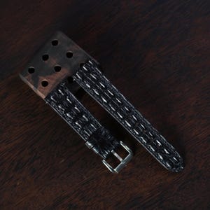 Può includere: Un cinturino nero per orologio con motivo a coccodrillo e fibbia argentata. Il cinturino è attaccato a una base di legno scuro con sei fori. Il cinturino è esposto su una superficie di legno marrone scuro.