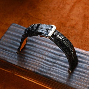 May include: A black alligator leather watch strap with a silver buckle. The strap has a textured finish and an orange underside. The watch band is displayed on a dark wooden surface against a brown backdrop.