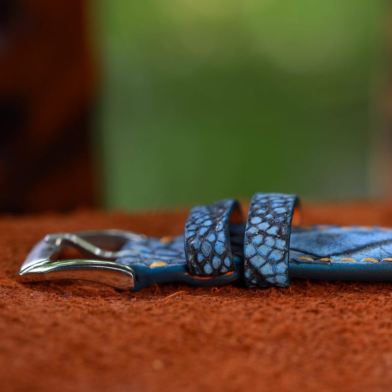 May include: Close-up of a blue watch strap with a textured pattern and a silver buckle. The strap has orange stitching and a detailed, scale-like design. The background is blurred, with a focus on the watch strap.