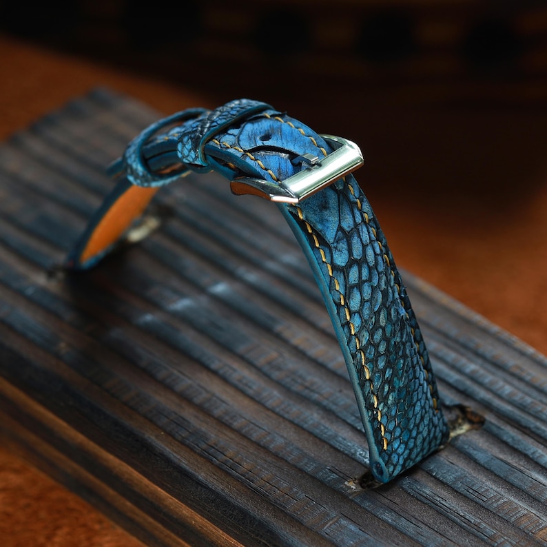 May include: A blue leather watch strap with a textured, reptile-like pattern. The strap features gold stitching and a silver-colored buckle. The watch band is displayed on a dark wooden surface.