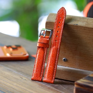 Puede incluir: Un par de correas de reloj de color naranja brillante con una superficie texturizada y costuras blancas. Las correas tienen una hebilla plateada y se muestran sobre una superficie de madera. Accesorio de reloj.