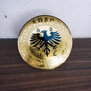 Può includere: Un emblema circolare in metallo color oro con un'aquila nera al centro. Il testo ADAC è stampato in alto, con le lettere A, D, A e C posizionate attorno all'aquila. Il bordo è decorato con un motivo a foglie di alloro in rilievo.
