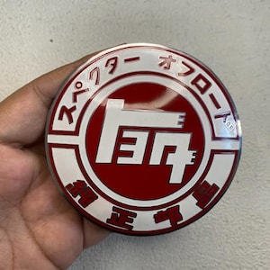 May include: A round, red and white emblem with Japanese text and the Toyota logo in white. The emblem has a metallic sheen and is likely a car part or accessory.
