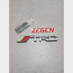 Könnte beinhalten: Ein silbernes TRD-Emblem mit roten und gelben Akzenten, zusammen mit einem ZEGEn-Aufkleber und einer kleinen Tüte mit Hardware. Das TRD-Logo ist ein stilisiertes Design.