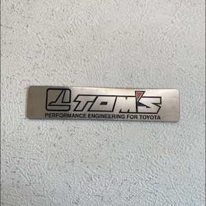 Puede incluir: Un emblema rectangular de metal plateado con el texto "TOMS" en una fuente audaz y estilizada. Debajo, se lee "PERFORMANCE ENGINEERING FOR TOYOTA". El emblema tiene un acento rojo.