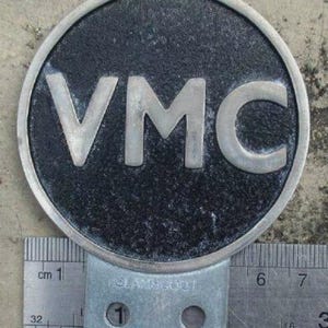 Puede incluir: Un emblema metálico redondo, gris oscuro, con las letras "VMC" en plata. El emblema tiene un borde plateado y se muestra con una regla para la escala. La regla está marcada en centímetros.