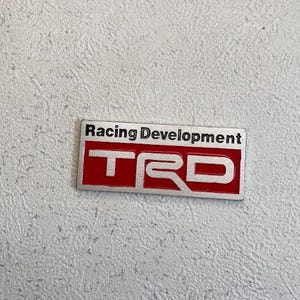 May include: A rectangular TRD emblem with the words "Racing Development" above the letters "TRD". The letters are silver with a red background and a black border. The emblem is on a textured, light grey surface.