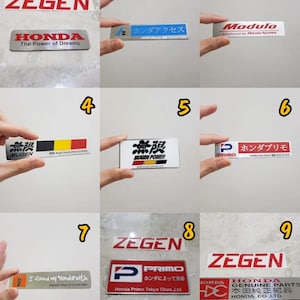 May include: A collection of car emblems and badges, featuring various designs and text. The emblems include the Honda logo, the text "ZEGEN", "MUGEN", and "PRIMO". Some badges have Japanese text and colorful accents.
