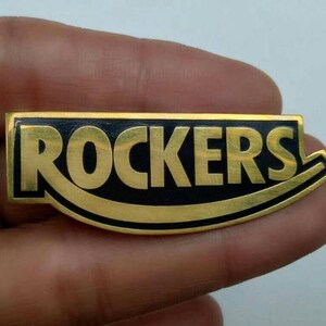 Może przedstawiać: Prostokątna przypinka z napisem "ROCKERS" w złotych literach na czarnym tle. Przypinka ma złotą obwódkę i zakrzywiony element. Przypinka jest trzymana w dłoni.