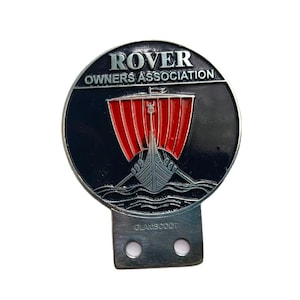Può includere: Un emblema metallico circolare con le parole "ROVER OWNERS ASSOCIATION" in alto. L'emblema presenta una nave vichinga con vele rosse su sfondo nero. La parte inferiore riporta la scritta "GLAMSCOOT".