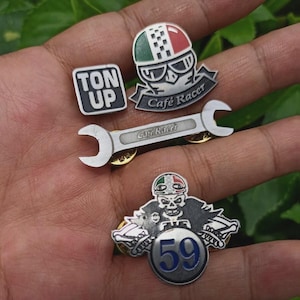 Puede incluir: Colección de pines metálicos plateados con temática de motociclismo. Los diseños incluyen una llave inglesa, una calavera con casco, un casco y las palabras "Ton Up" y "Cafe Racer". Los pines tienen detalles en rojo, blanco, verde y azul.