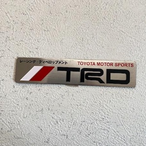 Könnte beinhalten: Ein silbernes TRD-Emblem mit den Worten "TOYOTA MOTOR SPORTS" in Rot. Das Emblem hat ein weißes und rotes Streifendesign auf der linken Seite und japanischen Text über dem TRD-Logo.