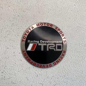 May include: A circular metal emblem with the text "TOYOTA MOTOR SPORTS RACING DEVELOPMENT" in red and silver. The center features "TRD" in white and red on a black background.