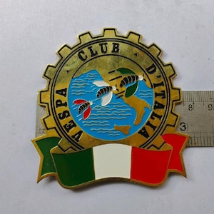 Puede incluir: Un emblema dorado del Vespa Club D'Italia con un borde en forma de engranaje. El emblema presenta un mapa de Italia, tres abejas estilizadas y la bandera italiana. El texto "VESPA CLUB D'ITALIA" está inscrito alrededor del mapa.
