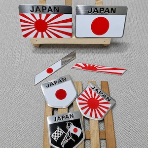 Może przedstawiać: Kolekcja siedmiu metalowych emblematów z napisem JAPAN czarną czcionką. Wzory obejmują japońską flagę wschodzącego słońca, flagę narodową z czerwonym kołem oraz emblemat w kształcie tarczy z szachownicą i mapą Japonii.