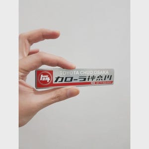 May include: A rectangular silver and red Toyota emblem with Japanese text and the words "TOYOTA CHUO OSAKA". The emblem is held between fingers.