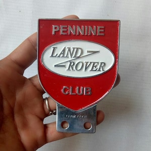 Peut inclure: Un emblème rouge et argenté du Pennine Land Rover Club. De forme de bouclier, il affiche les mots « PENNINE » et « CLUB ». Le logo Land Rover est au centre, dans un ovale.