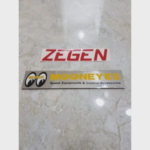 May include: Two rectangular stickers on a light gray surface. The top sticker has the word "ZEGEN" in red block letters. The bottom sticker has the "MOONEYES" logo with yellow text that reads "MOONEYES Speed Equipments & Custom Accessories."