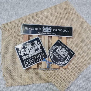 May include: Three black and silver emblems featuring the text Junction Produce and a crest with the letters JP. One rectangular emblem, one square emblem with rounded corners, and one shield-shaped emblem are arranged on wooden slats.