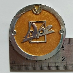 Puede incluir: Un emblema metálico redondo de color plateado con un centro dorado. El emblema presenta un logotipo estilizado con la palabra "Adoz" en plata. El emblema mide aproximadamente 5 cm de diámetro y tiene dos pequeños agujeros.