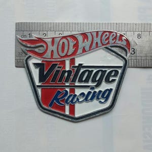 May include: A metal badge featuring the Hot Wheels logo in red and silver, with the words "Vintage Racing" in black and blue. The badge is approximately 7.6 cm long.
