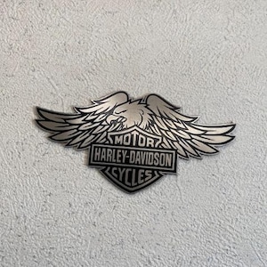 Könnte beinhalten: Silbernes Harley-Davidson-Emblem mit einem Adler mit ausgebreiteten Flügeln. Das Emblem enthält die Worte "MOTOR HARLEY-DAVIDSON CYCLES" in einem rechteckigen und schildförmigen Design.