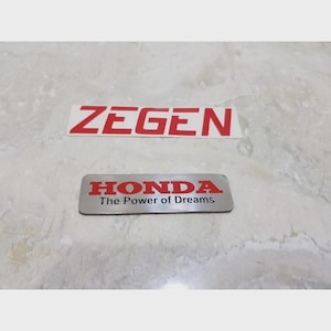 May include: Two automotive emblems. The first is a rectangular sticker with the word "ZEGEN" in red block letters. The second is a silver rectangular emblem with the word "HONDA" in red block letters, and the text "The Power of Dreams".