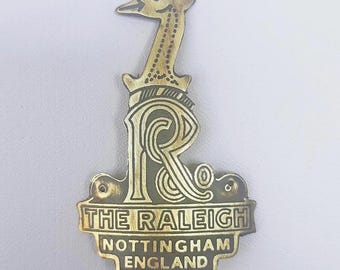 Könnte beinhalten: Ein messingfarbenes Metallabzeichen mit einem stilisierten Vogel auf einem dekorativen Buchstaben "R". Das Abzeichen enthält den Text "THE RALEIGH" über "NOTTINGHAM ENGLAND".