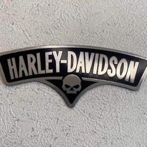 May include: A black and silver Harley-Davidson emblem with the brand name in silver lettering. The emblem features a skull design and is set against a textured background.